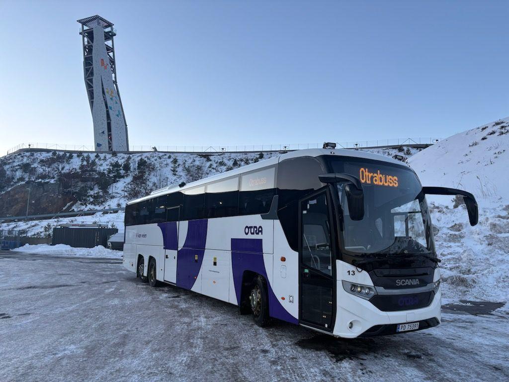 En moderne buss parkert i snøen med en skihoppbakke i bakgrunnen.