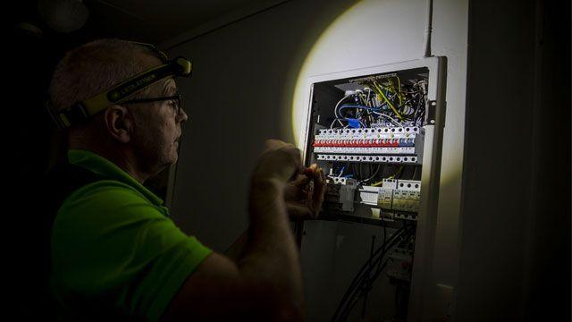 En elektriker jobber med en sikringsboks, opplyst av en hodelykt, med ledninger og komponenter synlige.