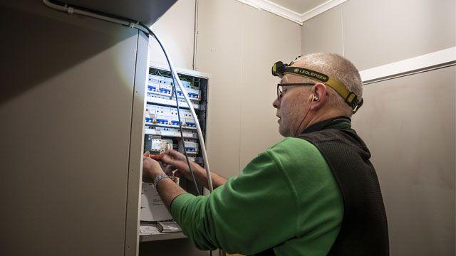 En elektriker jobber med sikringsskapet, iført hodelykt og headset, i et rom med grå vegger.