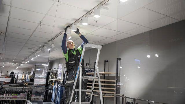 En arbeidstaker står på en stige og installerer belysning i en butikk.