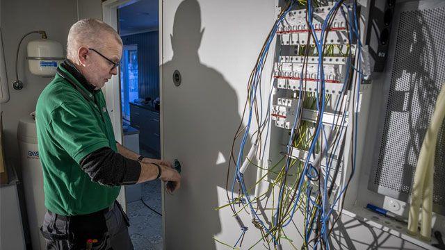En elektriker jobber med ledninger ved en dør, med elektriske komponenter synlige på veggen.