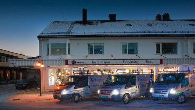 En bygning med butikk og tre varebiler parkert foran, under en klar himmel ved skumring.