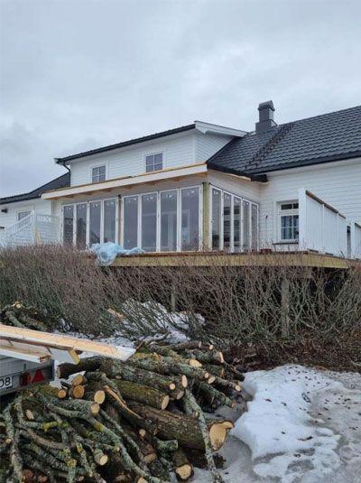 Hus med veranda og glassvegger, omringet av vedstabler og snø på bakken. Grå himmel i bakgrunnen.