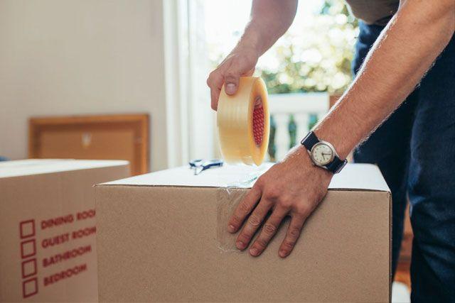 En person pakker en eske med tape, med fokus på hånden og esken i et lyst rom.