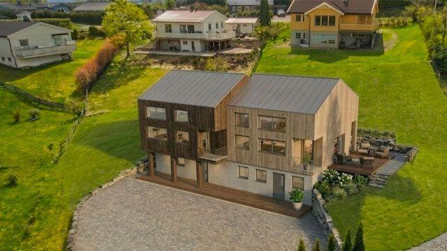 Moderne hus med treverk og store vinduer, omgitt av grønt gress og hager i et boligområde.