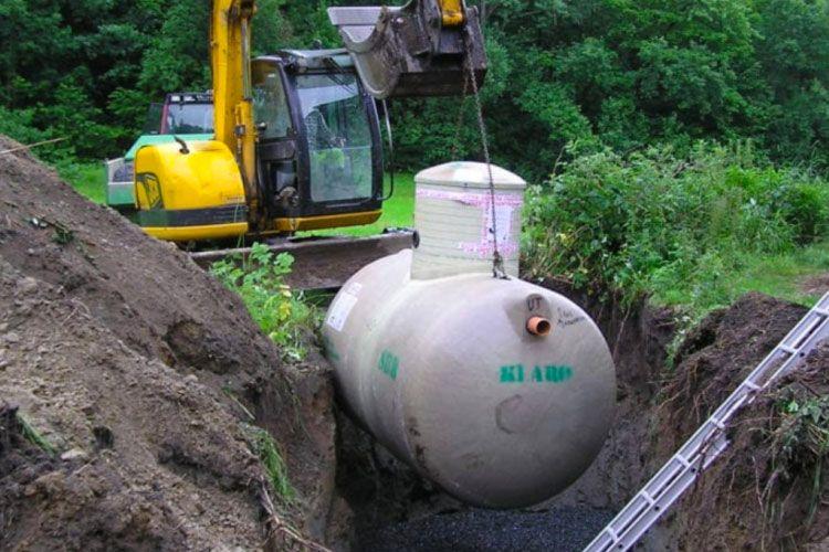 Gravemaskin heiser en stor tank opp fra en grøft i en grønn hage.