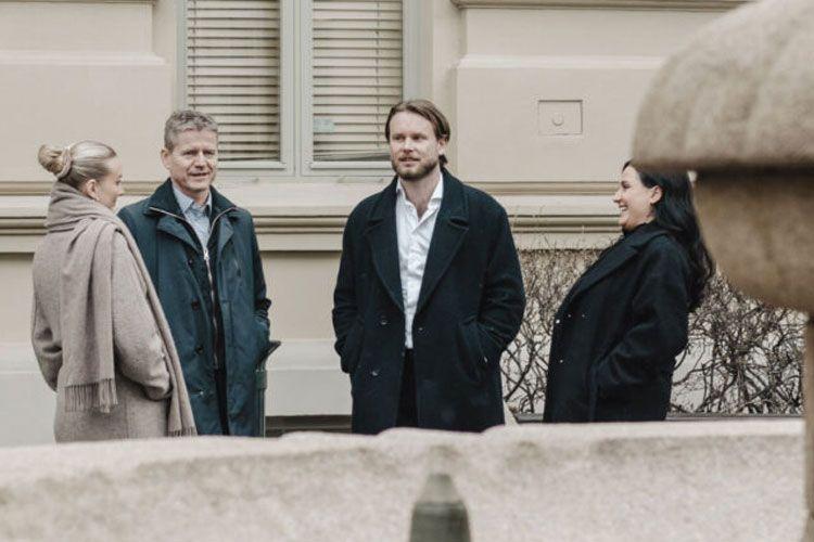 Fire personer står sammen ved en fontene, iført vinterklær, med bygninger i bakgrunnen.