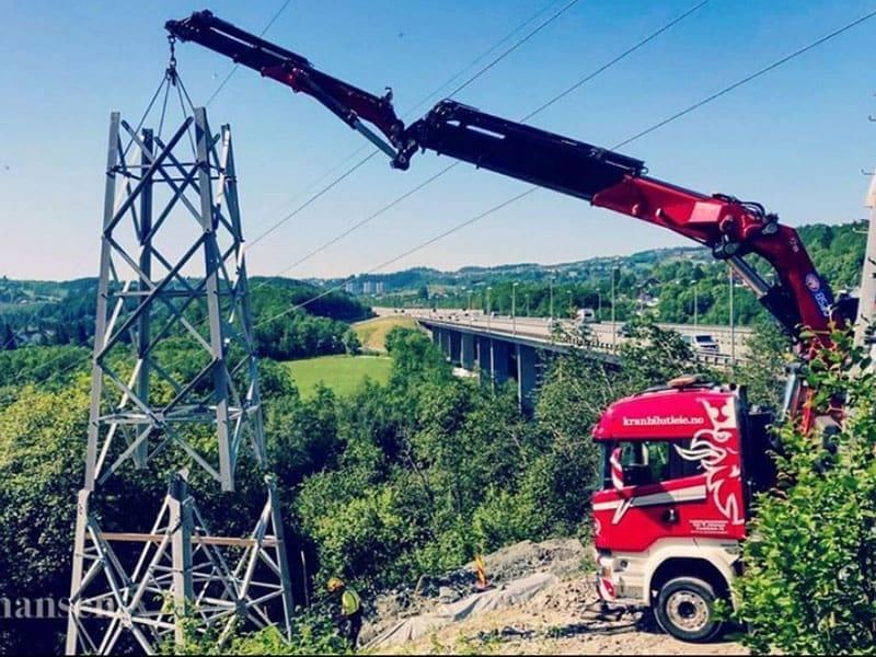 En lastebil med kran heiser en høyspentmast i et grønt landskap med en bro i bakgrunnen.