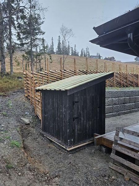 Liten svart bod med skråtak ved en gjerde i et skogsområde, omringet av trær og åpen mark.