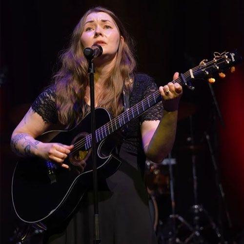 Kvinne synger med gitar på scenen, iført svart kjole og med mikrofon foran seg.