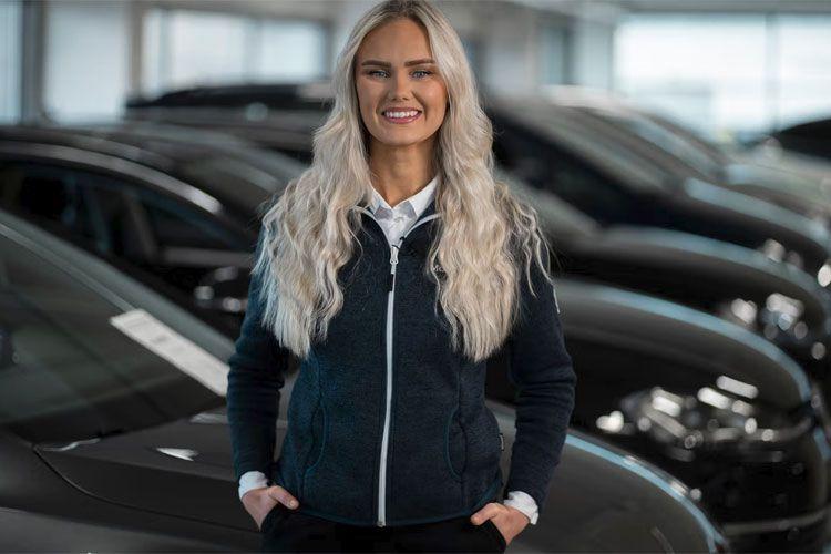 Kvinne med langt, lyst hår står smilende foran svarte biler i en bilforhandler.