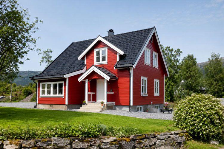Rødt hus med skråtak og hvite vinduer, omgitt av grønt gress og trær i et naturskjønt landskap.
