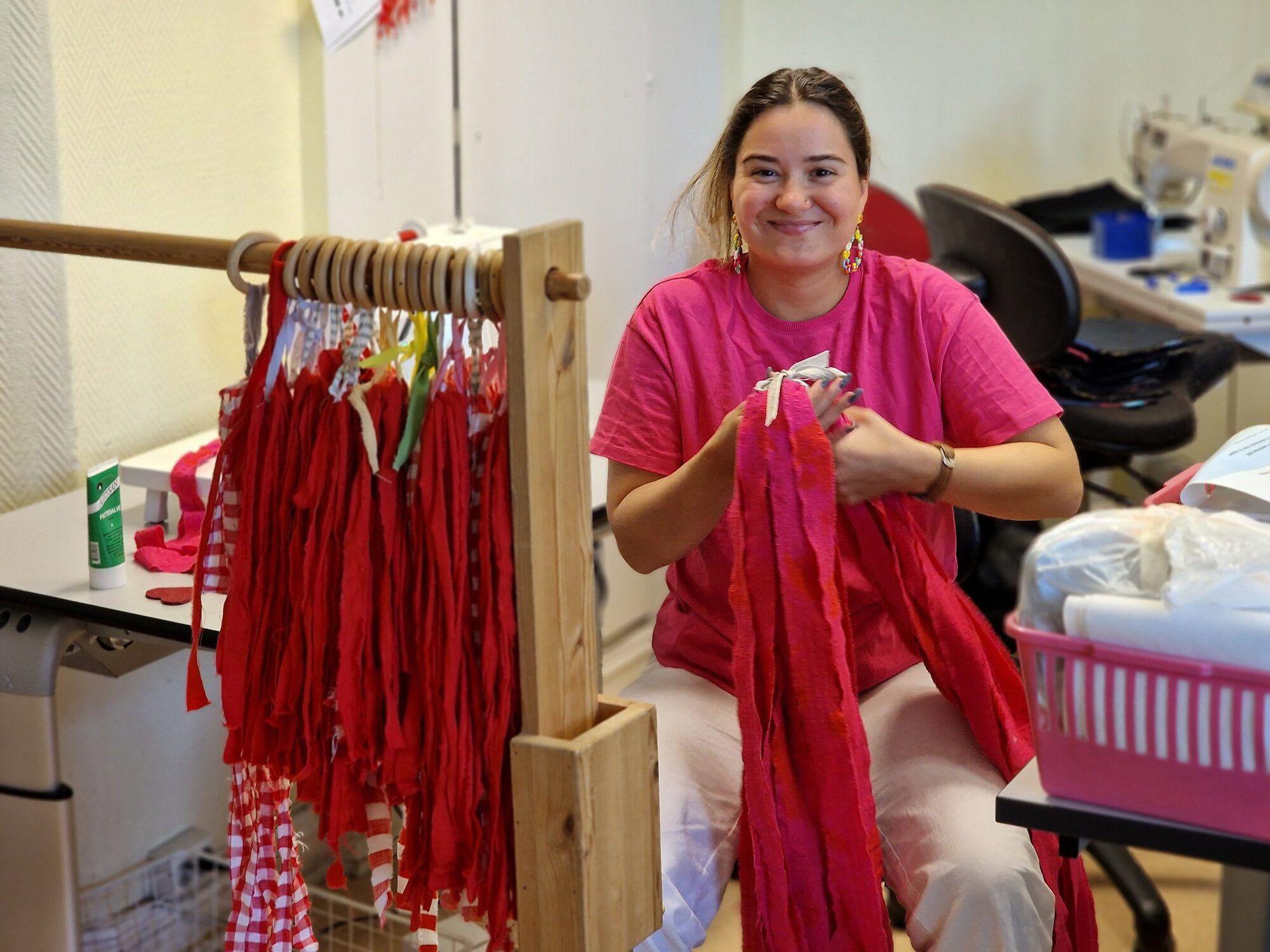 Kvinne i rosa t-skjorte som lager røde bånd ved en arbeidsbenk med syutstyr.