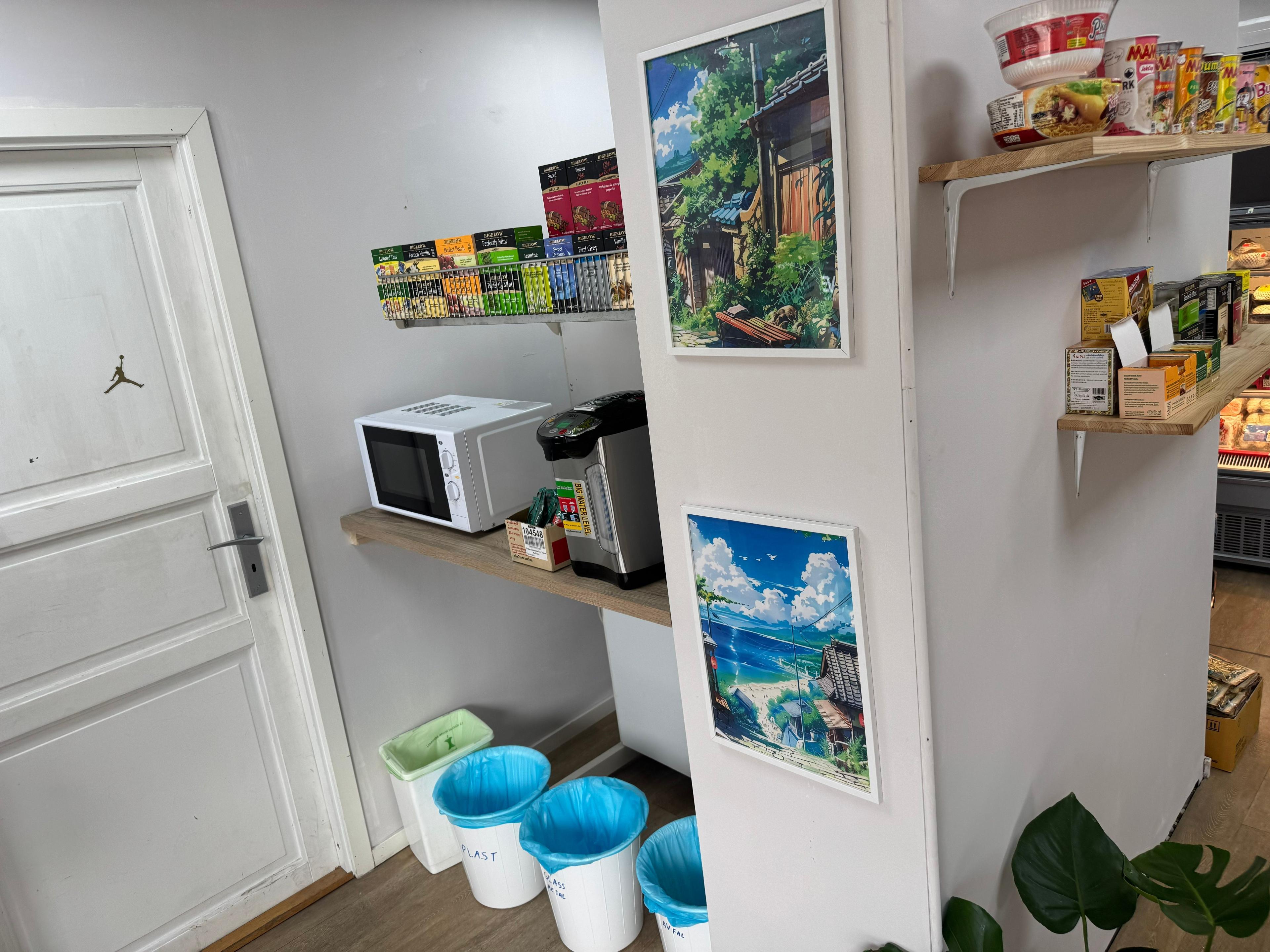 Kjøkkenhjørne med mikrobølgeovn, kaffemaskin, fargerike bilder på veggen og søppelbøtter i hjørnet.