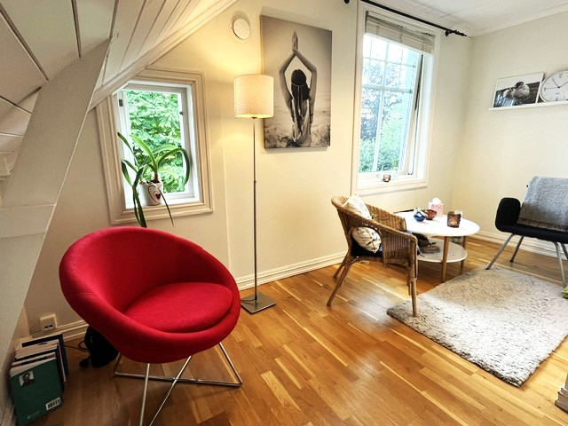 Lyst stue med rød stol, hvit lampe, planter, og et bord med bøker og dekorasjoner. Veggen har et kunstverk.
