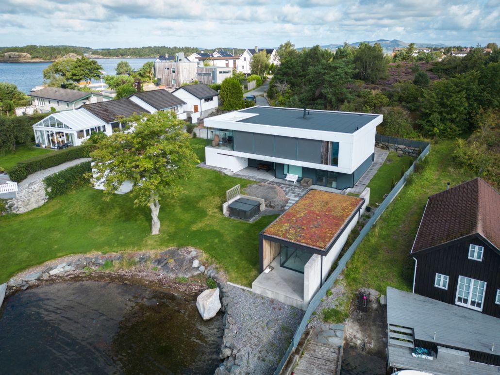 Moderne hus med flatt tak og stor hage ved vannet, omgitt av trær og andre boliger.