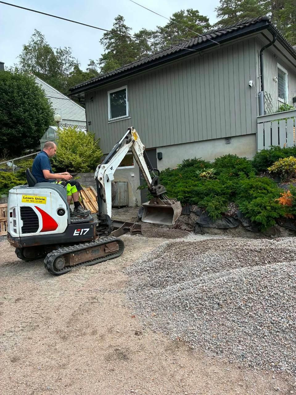 En mann som kjører en gravemaskin på en gruslagt hage foran et hus med busker og trær.