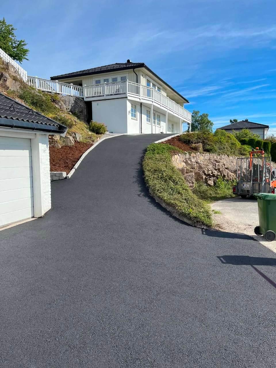 Moderne hus med balkong, beliggende på en skråning med asfaltert innkjørsel og grønt landskap rundt.