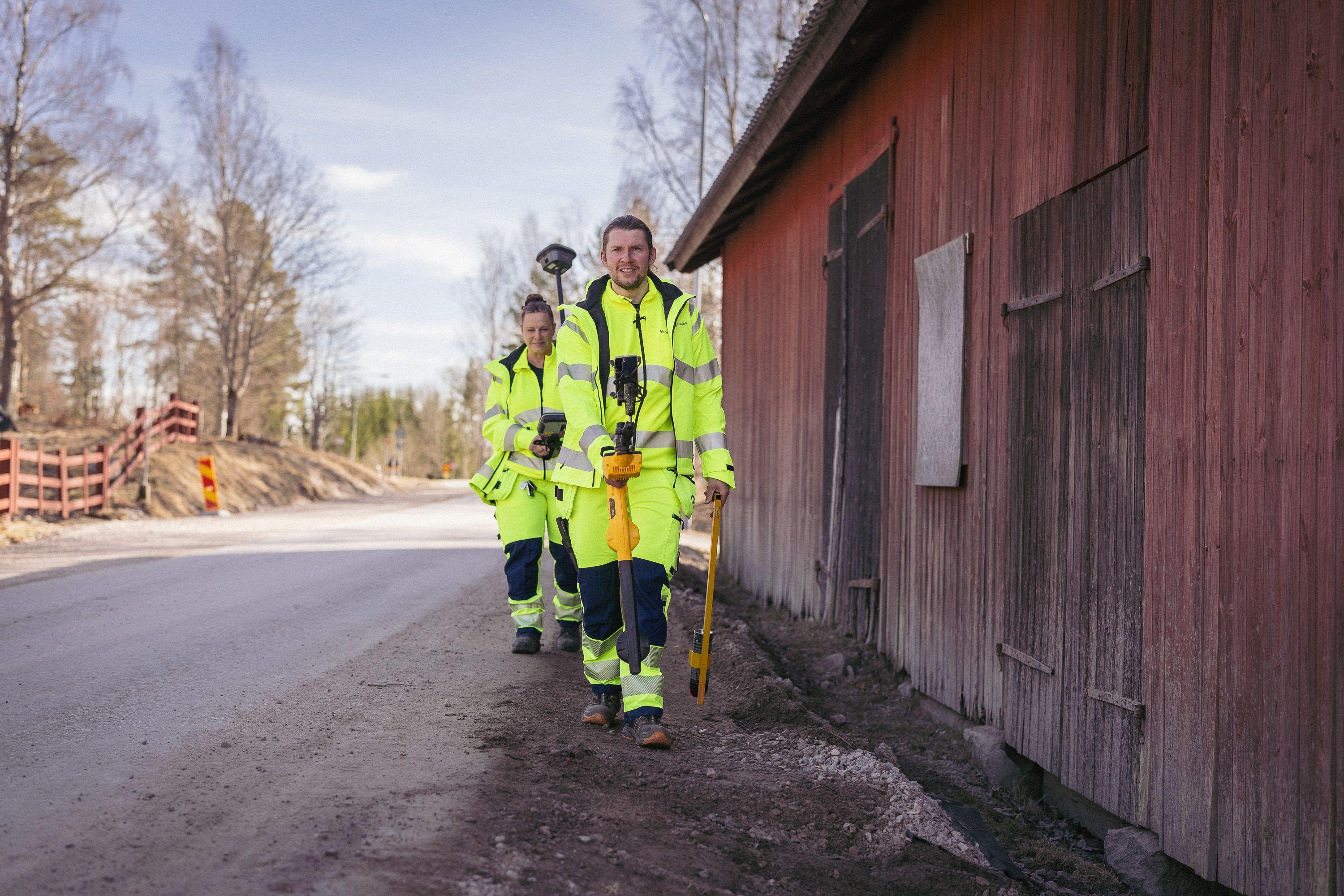 To personer i refleksjakker går langs en vei ved en rød bygning, med verktøy i hånden.
