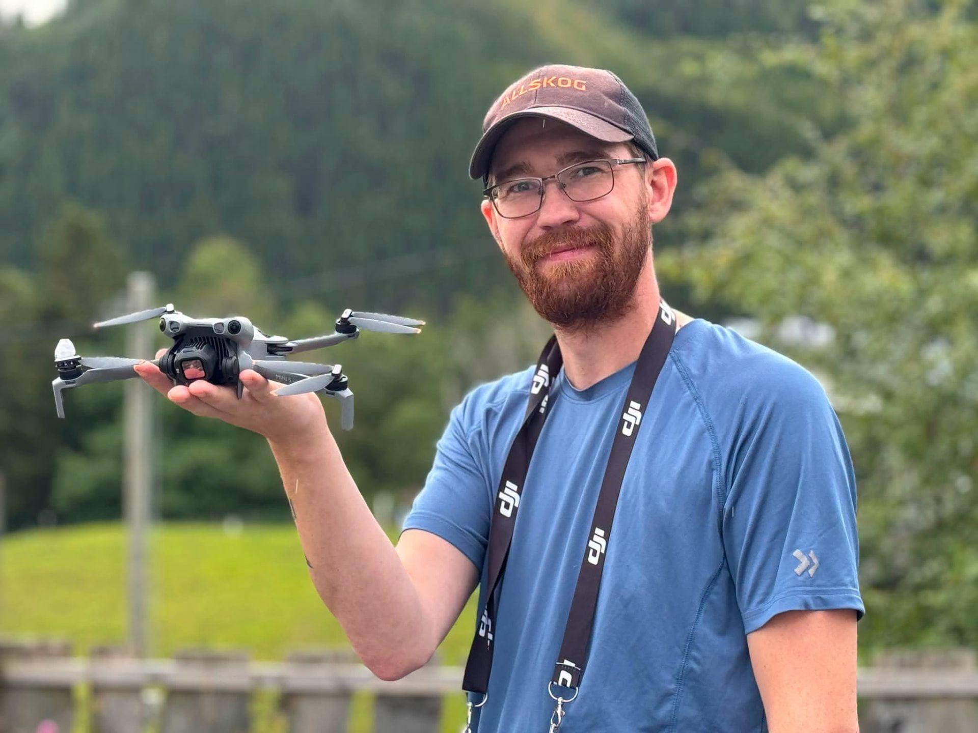 En mann med skjegg holder en drone i hånden, med grønne fjell i bakgrunnen.