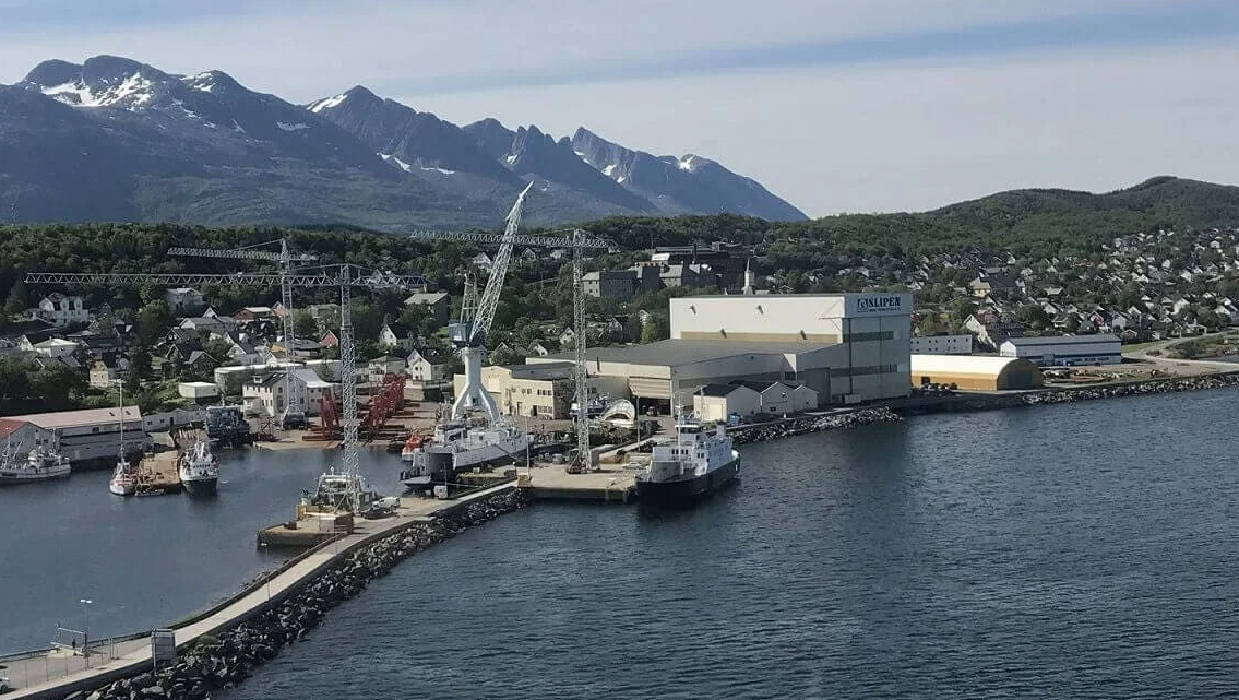 Havn med båter, kraner og industriområder, omgitt av fjell med snøtopper og grønn vegetasjon.