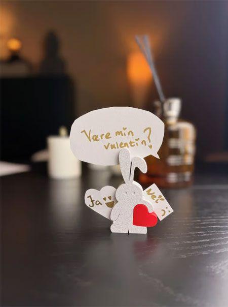 En papirkanin med hjerter og en snakkeboble som spør "Være min valentin?" på et bord.