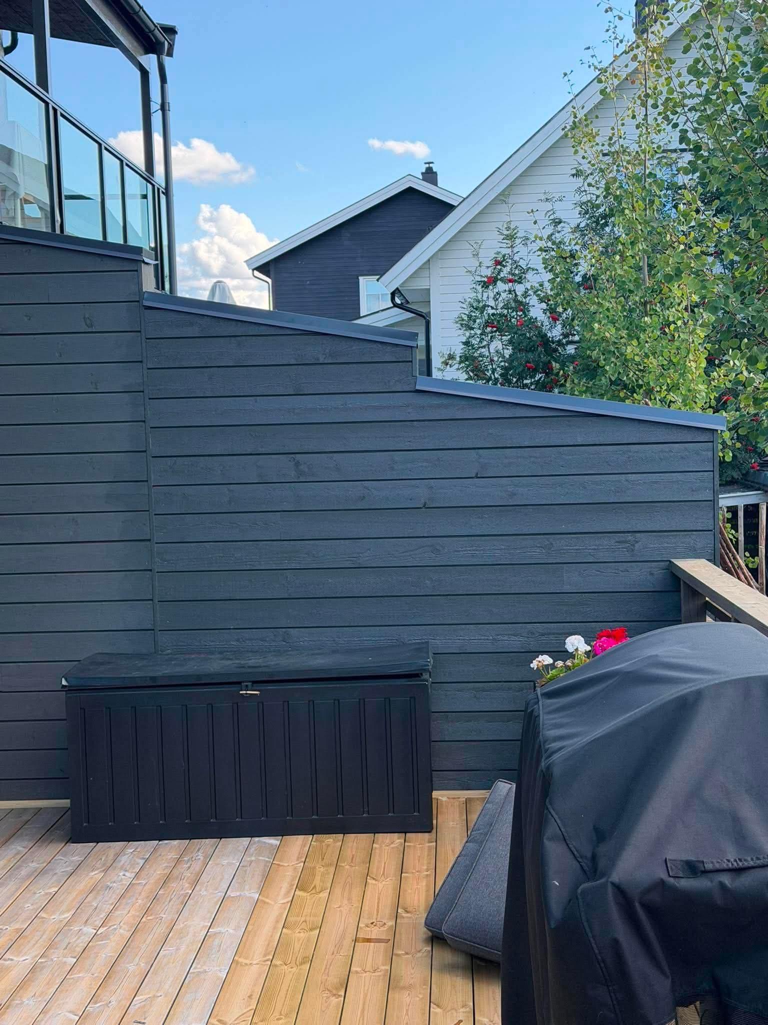 Utsikt over en terrasse med en svart oppbevaringsboks og en grill, omgitt av grønne trær og hus.