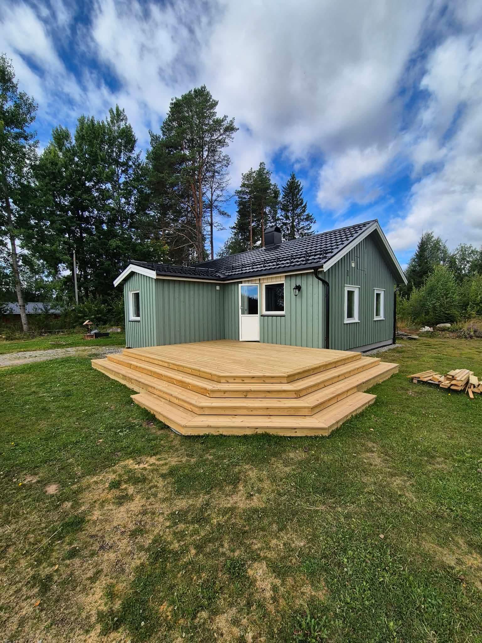 En moderne hytte med grønn fasade og terrasse i tre, omgitt av trær og blå himmel.