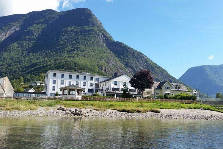 Utsikt over et hotell ved vannet, med fjell i bakgrunnen og grønt landskap rundt.
