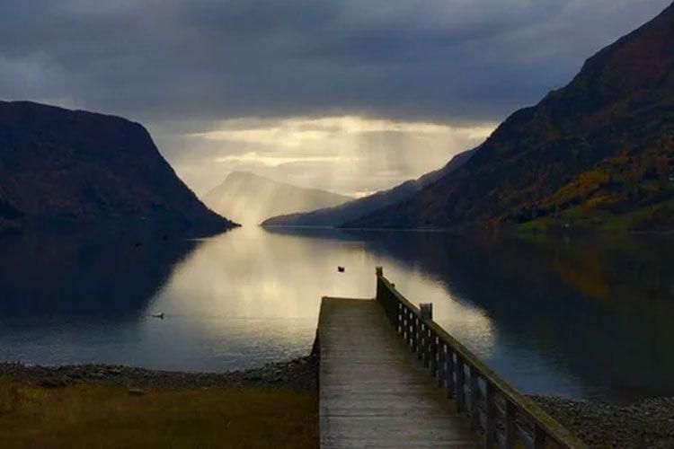 En vakker utsikt over en stille innsjø med fjell i bakgrunnen og solstråler som skinner gjennom skyene.