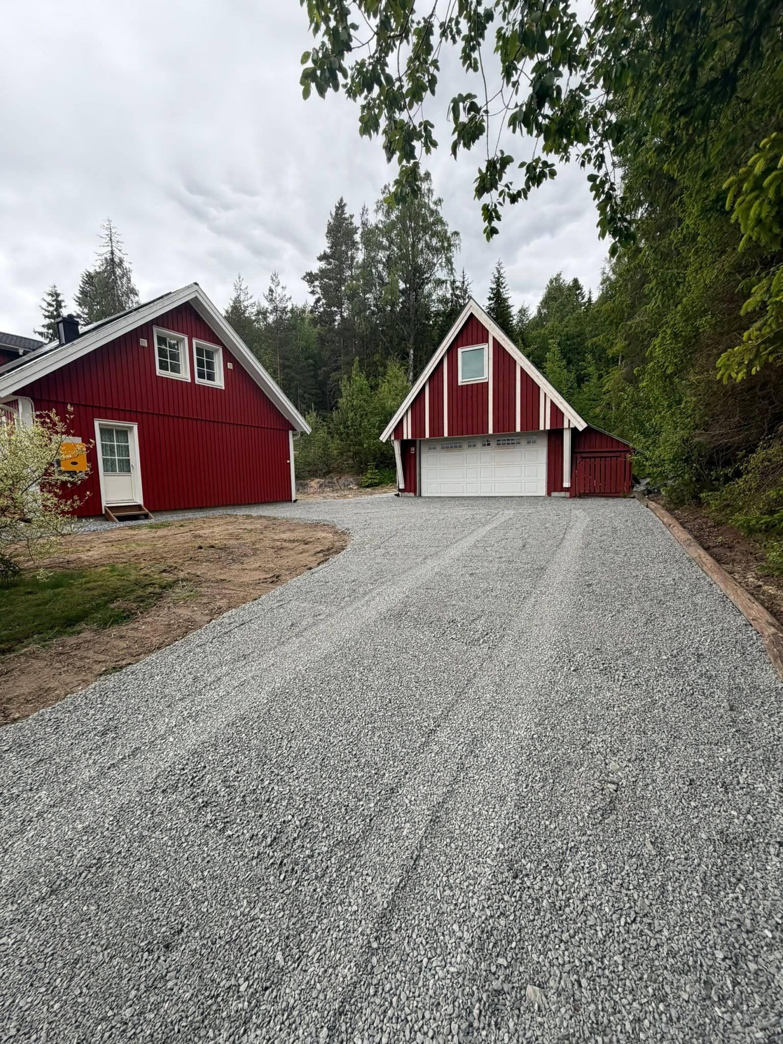 Rød hytte med garasje ved en gruslagt innkjørsel omringet av trær og grønt landskap.
