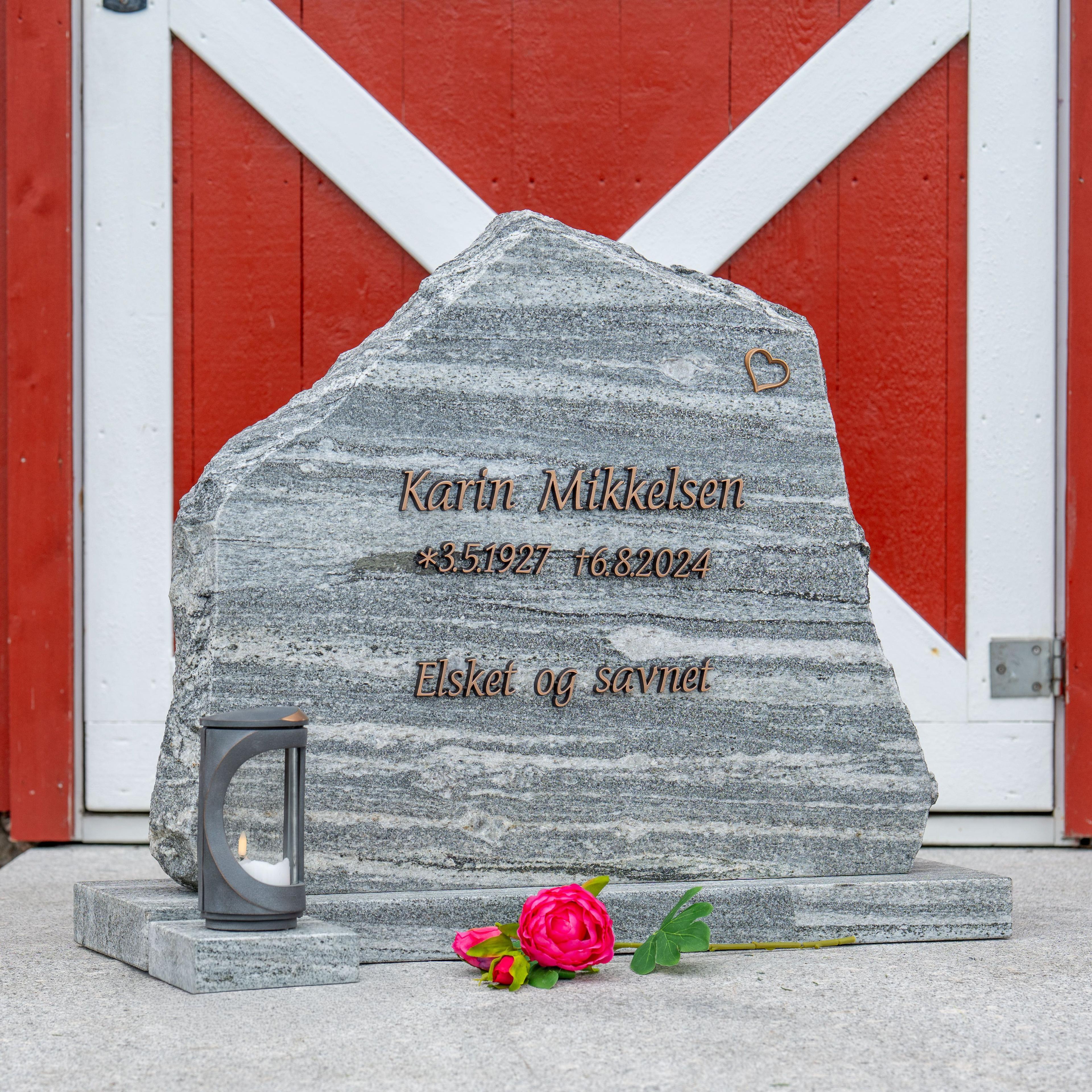 Grå gravstein med inskripsjon for Kåre Mikkelsen, med en lykt og en rose foran, plassert mot en rød bakgrunn.