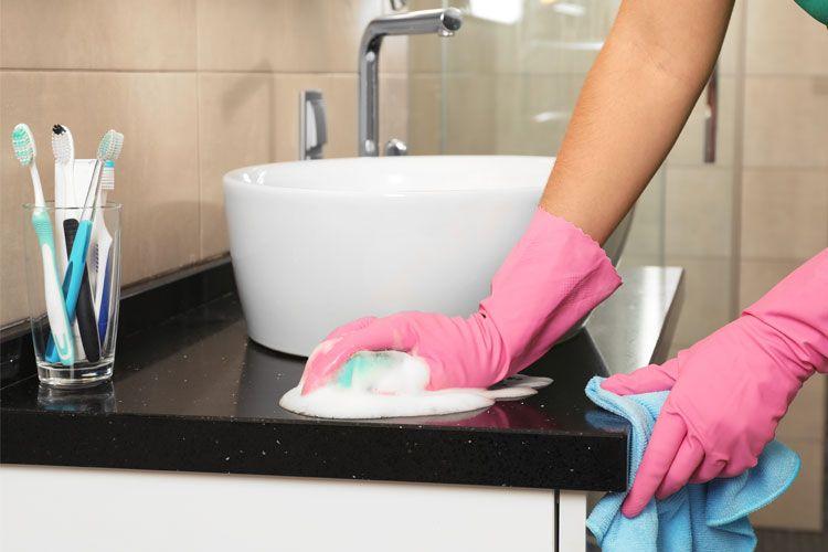 En person med rosa gummihansker vasker en benkeplate på badet med en klut og såpe.
