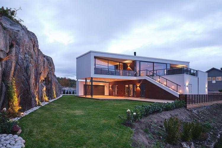 Moderne hus med store vinduer, beliggende ved en steinete skråning og grønt gressområde.