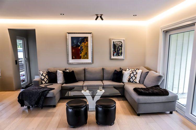 Moderne stue med grå sofa, glassbord, svarte puffer og kunstverk på veggen. Naturlig lys fra vinduet.