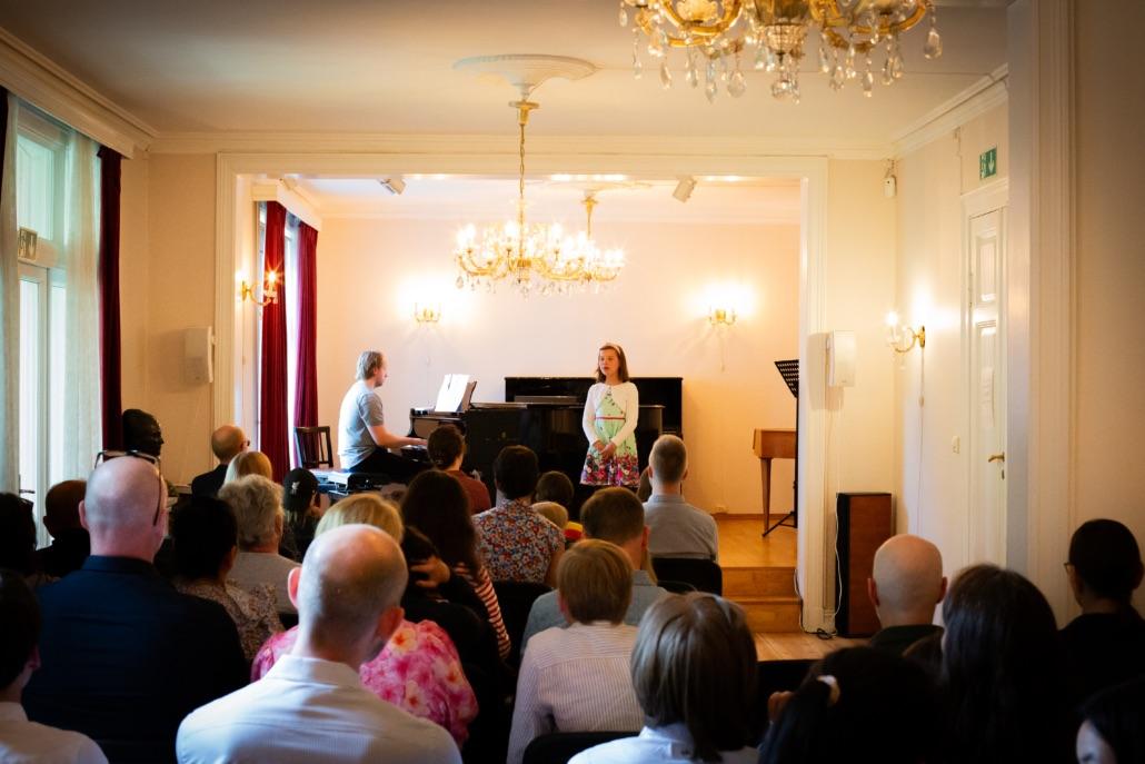 En konsert med en sangerinne som fremfører på scenen, mens en pianist spiller piano foran et publikum.