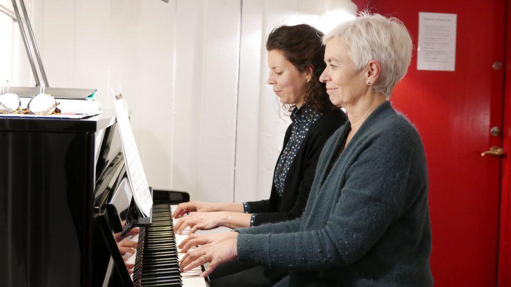 To kvinner som spiller piano sammen, med noteark foran dem i et lyst rom.
