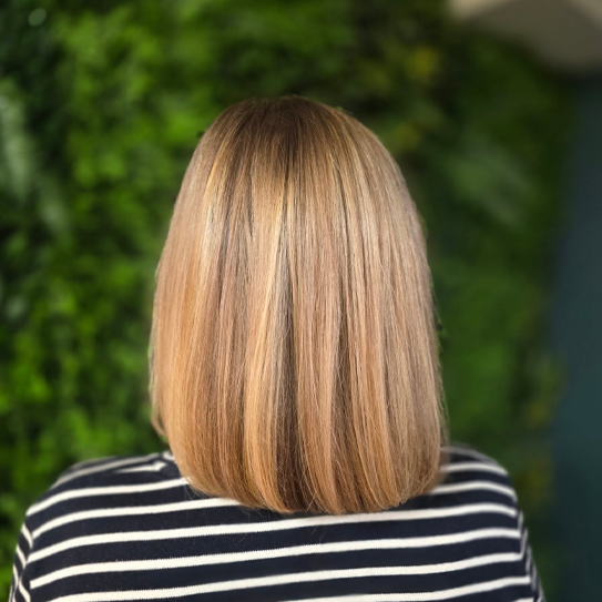 En kvinne med langt, rett hår i lys blond farge, sett bakfra mot en grønn bakgrunn.