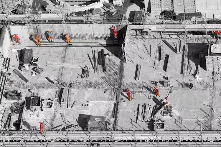 Byggeplass med arbeidere i oransje klær som jobber med betong og stål på en byggeflate.