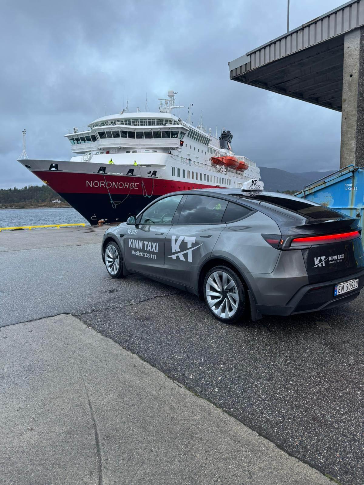 En grå taxi står parkert ved kaien med et stort ferje i bakgrunnen. Mørke skyer henger over området.