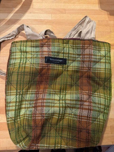 En rutete totebag i grønne og brune farger med lange håndtak og en logo på forsiden.