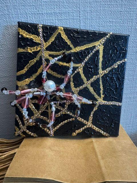 Et kunstverk med en svart bakgrunn, gull- og sølvfargede edderkoppnett og en dekorativ edderkopp laget av perler.