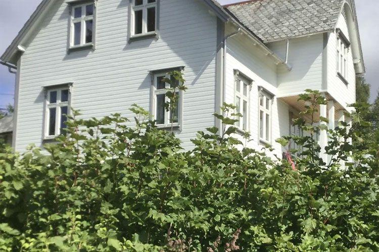 Hvit trehus med skråtak, omringet av grønne busker og planter i solfylt vær.