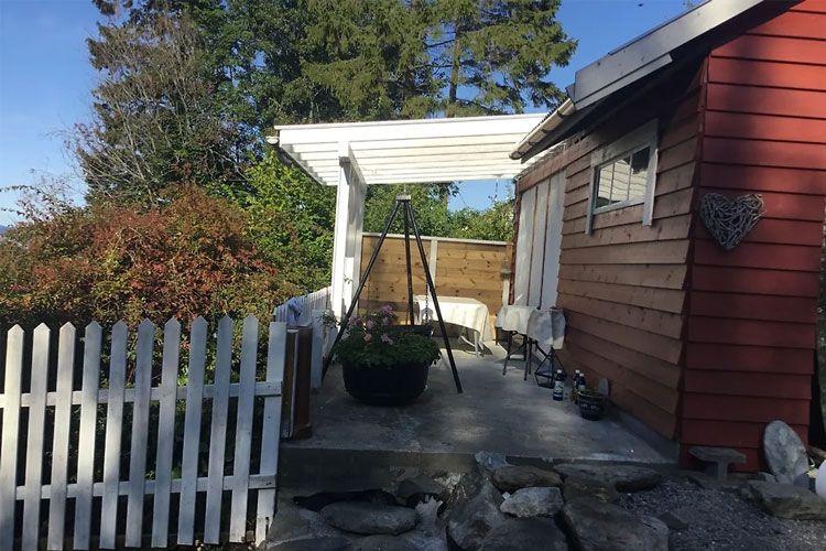 En koselig terrasse med hvit pergola, rød husvegg og hageplanter, omgitt av en hvit picket-gjerde.