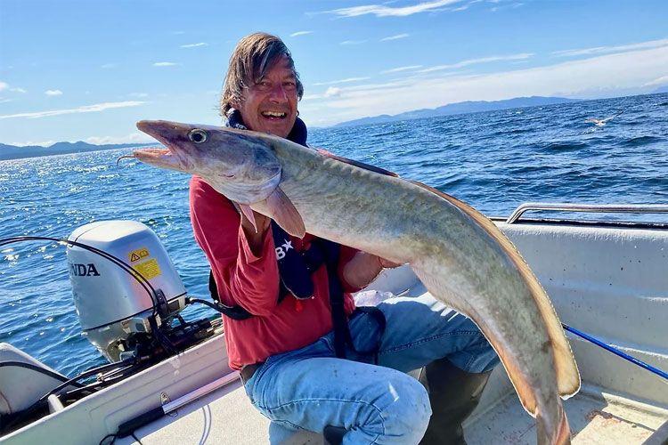 En mann sitter i en båt og holder en stor fisk mens han smiler mot kameraet. Havet er klart i bakgrunnen.