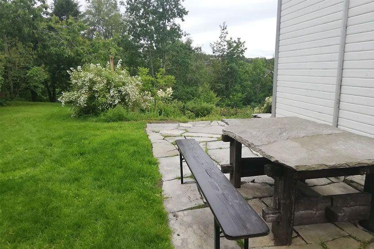 Uteområde med steinlagt terrasse, benk og bord, omkranset av grønt gress og busker.