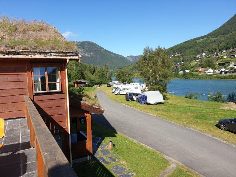 Utsikt fra en hytte med tak av gress, mot en elv og campingvognpark i fjellandskap.