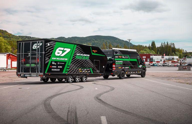 En svart og grønn racingbil med tilhenger på en parkeringsplass, omgitt av trær og bygninger i bakgrunnen.