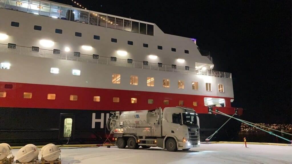 Lastbil fyller drivstoff på et cruiseskip i havn om natten, med lys som lyser opp skipets side.