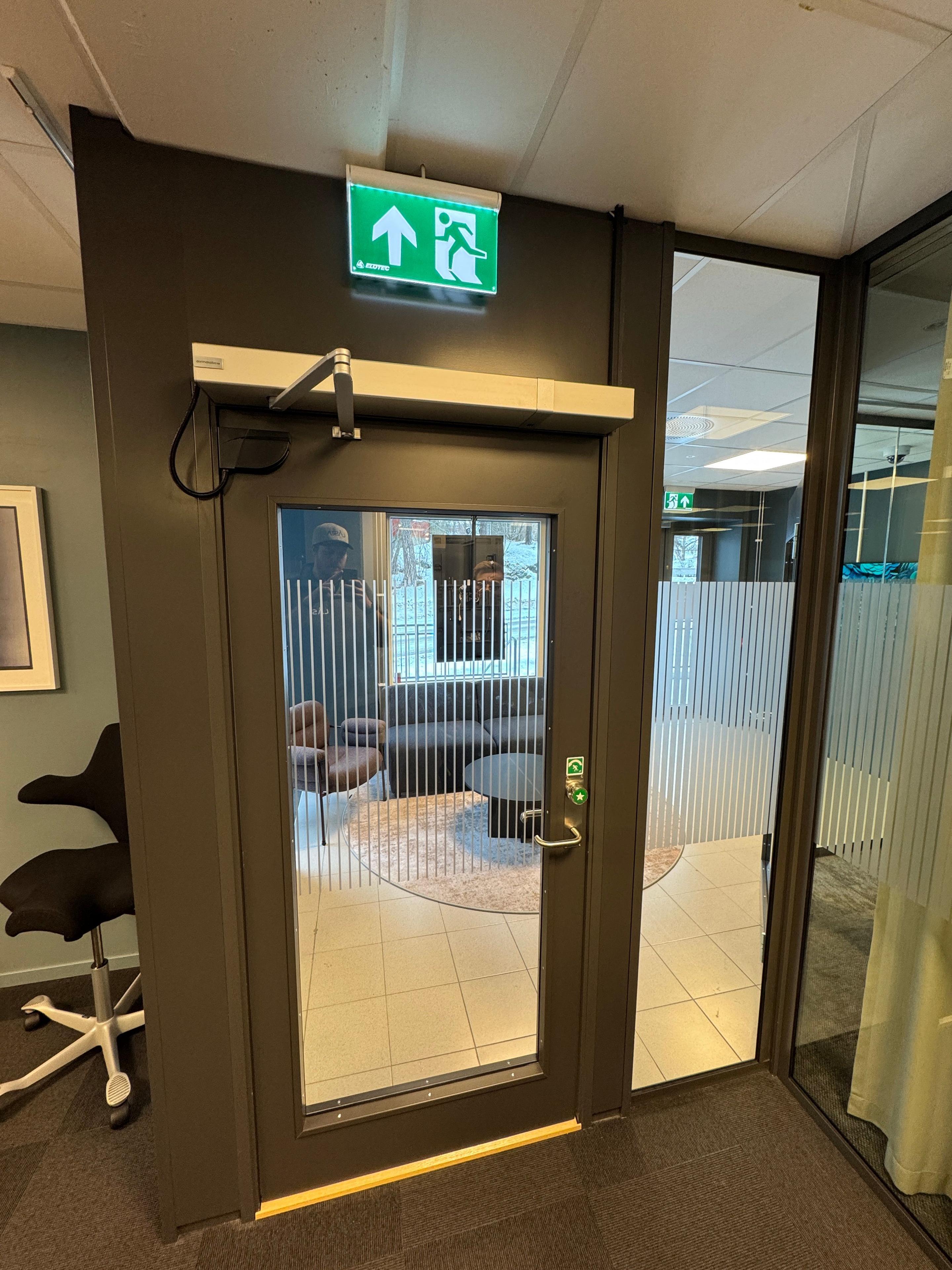 Dør med glassvindu og nødutgangssymbol, plassert i et moderne kontorlandskap.
